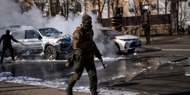 Soldados ucranianos toman posición frente a una instalación militar mientras dos autos se queman, en una calle de Kiev, Ucrania, el sábado 26 de febrero de 2022. Las tropas rusas irrumpieron en la capital ucraniana el sábado y estallaron las protestas callejeras cuando los funcionarios de la ciudad instaron a los residentes a refugiarse.  . 