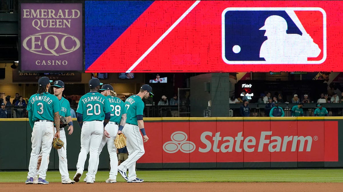 FILE - Seattle Mariners gather as the MLB logo is shown during a review of an attempted catch by right fielder Mitch Haniger of a ball hit by Tampa Bay Rays' Ji-Man Choi that was originally called an out during the ninth inning of a baseball game Friday, June 18, 2021, in Seattle. The call was overturned. While Major League Baseball and the players union try to chart a path forward, hoping to get baseball back on the field, some experts are watching the talks more from an academic viewpoint