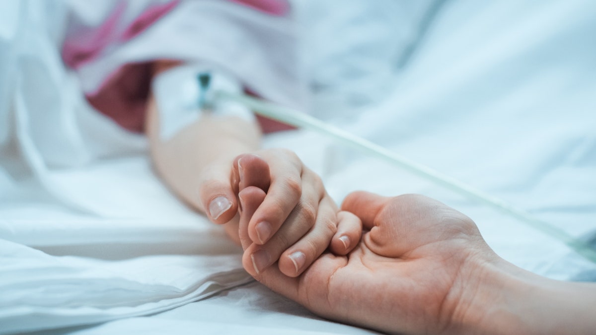 A child and parent hold hands in a hospital bed.