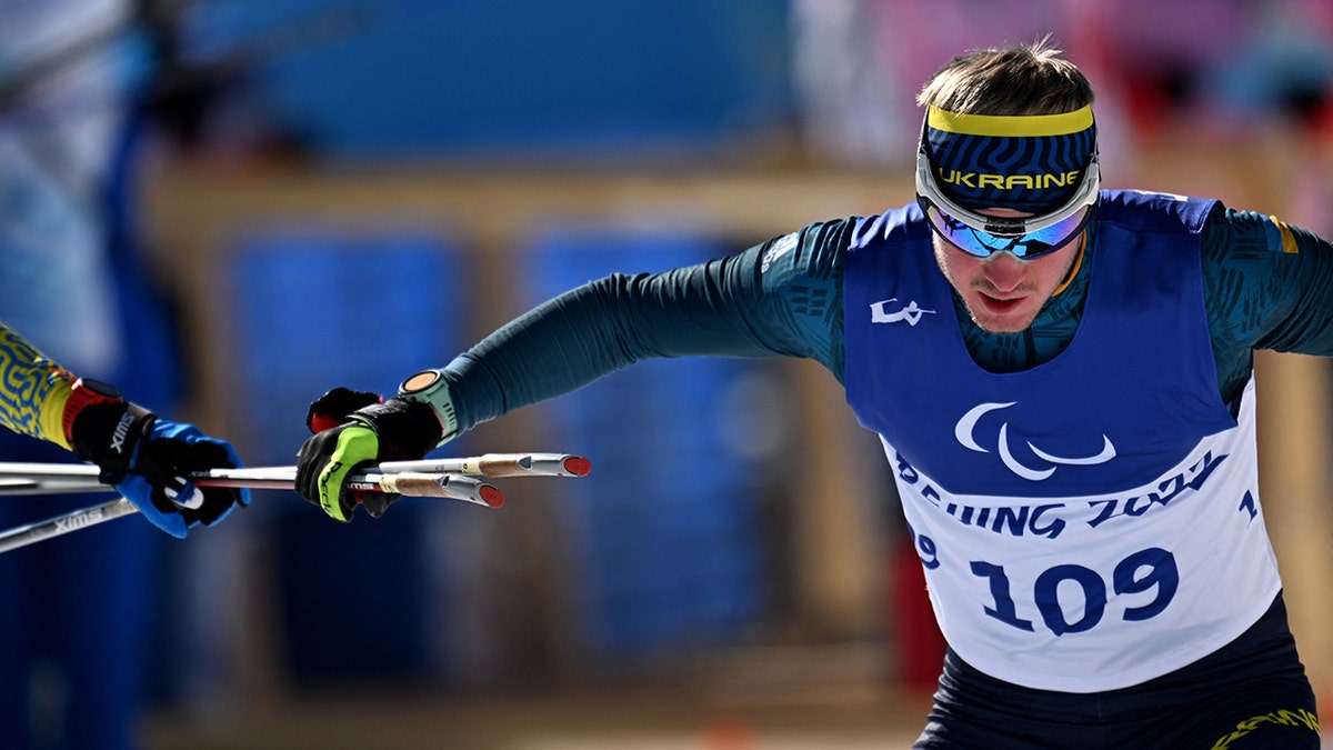 Ukraines Dmytro Suiarko grabs his poles from his guide in the mens sprint vision impaired biathlon event on March 5, 2022, at the Zhangjiakou National Biathlon Centre, during the Beijing 2022 Winter Paralympic Games.