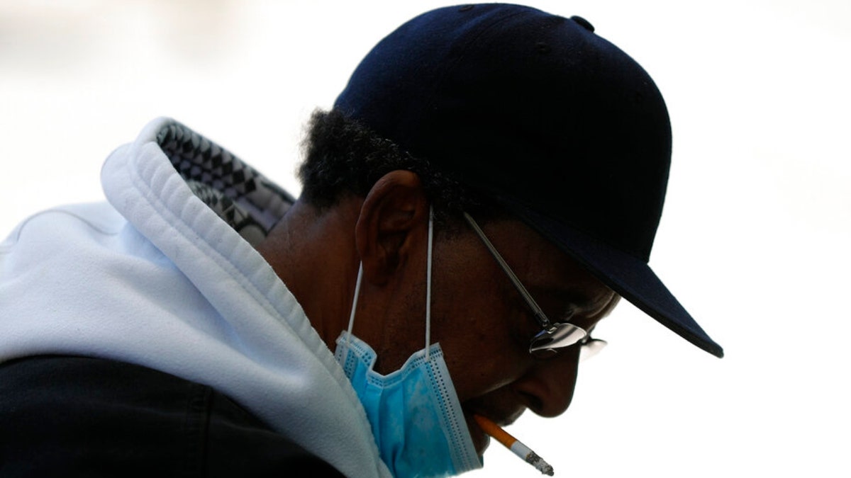 A man smokes a cigarette while wearing a protective mask while waiting for a bus in Detroit, Wednesday, April 8, 2020.