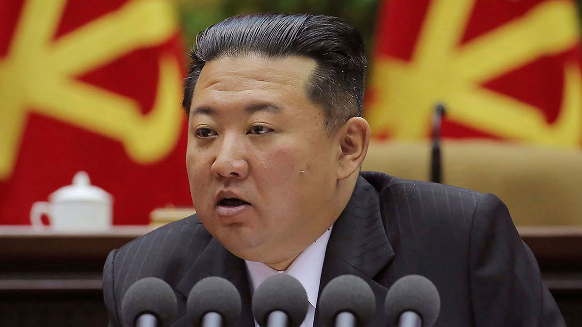 Kim Jong Un speaks into several mics while sitting in front of a red and yellow background