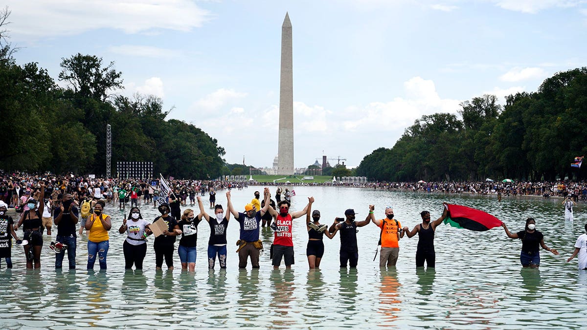 FILE - People join hands as they pose for a photo in the Reflecting Pool in the shadow of the Washington Monument as they attend the March on Washington, at the Lincoln Memorial in Washington, on the 57th anniversary of the Rev. Martin Luther King Jr.'s "I Have a Dream" speech on Friday, Aug. 28, 2020. California's first-in-the-nation task force on reparations is at a crossroads with members divided on which Black Americans should be eligible for compensation. The task force could vote on the question of eligibility on Tuesday, March 28, 2022, after putting it off at last month's meeting. (AP Photo/Julio Cortez, File)