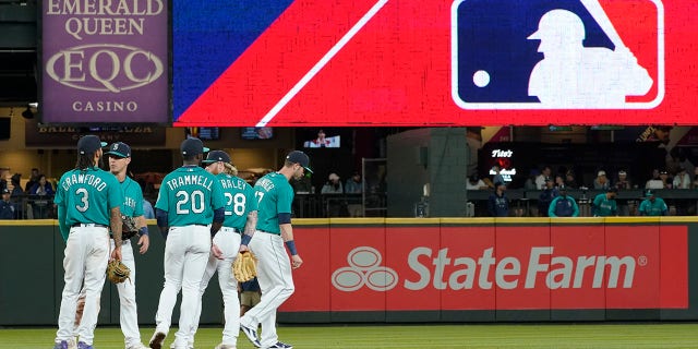 FILE - Seattle Mariners gather as the MLB logo is shown during a review of an attempted catch by right fielder Mitch Haniger of a ball hit by Tampa Bay Rays' Ji-Man Choi that was originally called an out during the ninth inning of a baseball game Friday, June 18, 2021, in Seattle. The call was overturned. While Major League Baseball and the players union try to chart a path forward, hoping to get baseball back on the field, some experts are watching the talks more from an academic viewpoint
