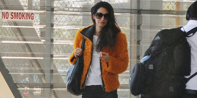 Amal Clooney dons a fashionable orange jacket, white t-shirt, dark trousers, and matching heels while at a Washington D.C. airport.