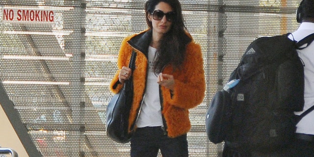 Amal Clooney dons a fashionable orange jacket, white t-shirt, dark trousers, and matching heels while at a Washington D.C. airport.