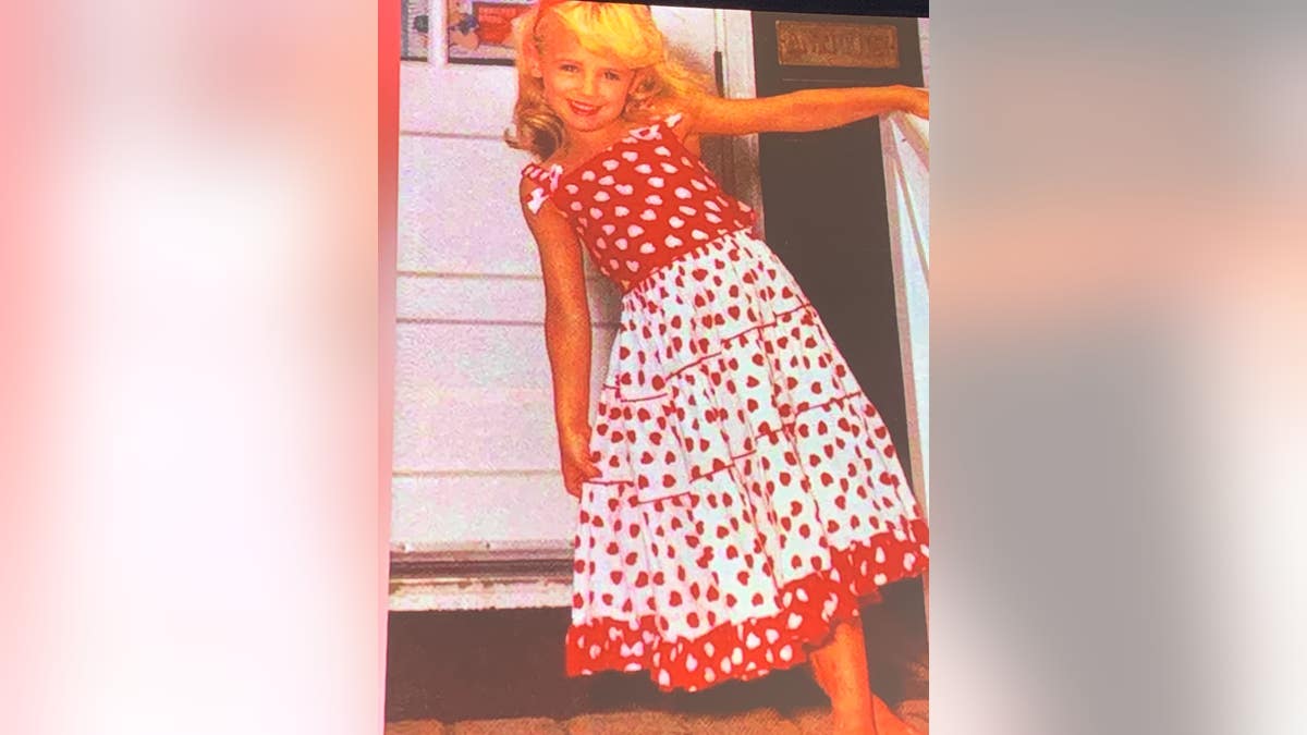 JonBenet Ramsey leans from a railing, smiling and wearing a red and white polka dot dress