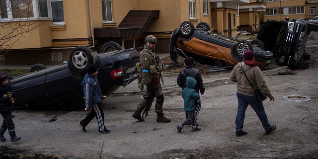 A Ukrainian soldier walks with children passing destroyed cars due to the war against Russia, in Bucha, on the outskirts of Kyiv, Ukraine, Monday, April 4, 2022. 