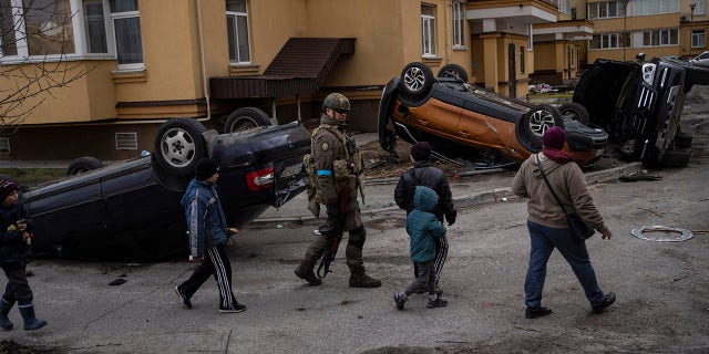 A Ukrainian soldier walks with children passing destroyed cars due to the war against Russia, in Bucha, on the outskirts of Kyiv, Ukraine, Monday, April 4, 2022.