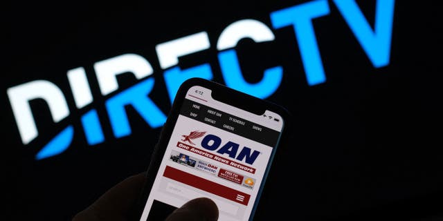 This illustration photo shows a person reading the One America Network website on a smartphone in front of a DirecTV logo in Los Angeles, January 14, 2022.