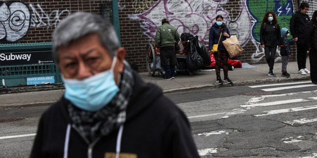 A man, wearing a protective face mask, walks by as others wait on a street corner where a man sleeps on a sidewalk, outside a subway station, in the Queens borough of New York City, U.S., April 18, 2022. 