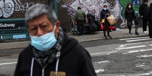 A man, wearing a protective face mask, walks by as others wait on a street corner where a man sleeps on a sidewalk, outside a subway station, in the Queens borough of New York City, U.S., April 18, 2022. 