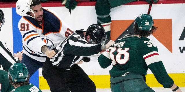 An official, center, gets caught between Edmonton Oilers' Evander Kane (91) and Minnesota Wild's Ryan Hartman (38) during a skirmish in the third period of an NHL hockey game Tuesday, April 12, 2022, in St. Paul, Minn. Both players received game misconduct penalties. The Wild won 5-1, with Hartman scoring two of the goals.