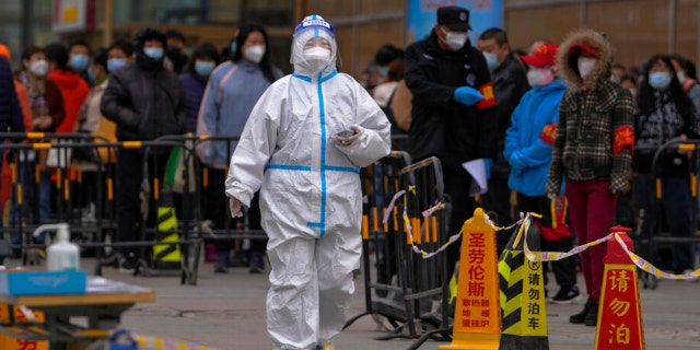 A health worker wearing a protective suit walks by masked residents who wait in line to get their throat swab at a coronavirus testing site after a COVID-19 case was detected in a residential building, Wednesday, April 6, 2022, in Beijing.