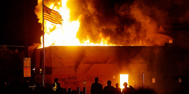KENOSHA, WI - AUGUST 24: People watch a the American flag flies over a burning building during a riot as demonstrators protest the police shooting of Jacob Blake on Monday, August 24, 2020 in Kenosha, Wisconsin. Blake was shot in the back multiple times by police officers responding to a domestic dispute call yesterday. 