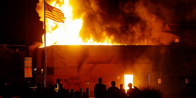 KENOSHA, WI - AUGUST 24: People watch the American flag flies over a burning building during a riot as demonstrators protest the police shooting of Jacob Blake on Monday, August 24, 2020 in Kenosha, Wisconsin.