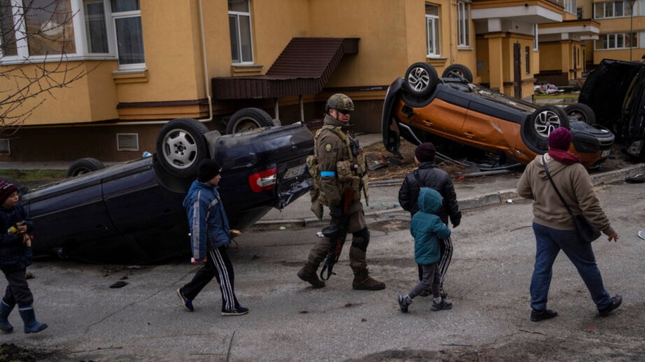 Ukrainian soldiers and kids walk by overturned cars in Bucha.