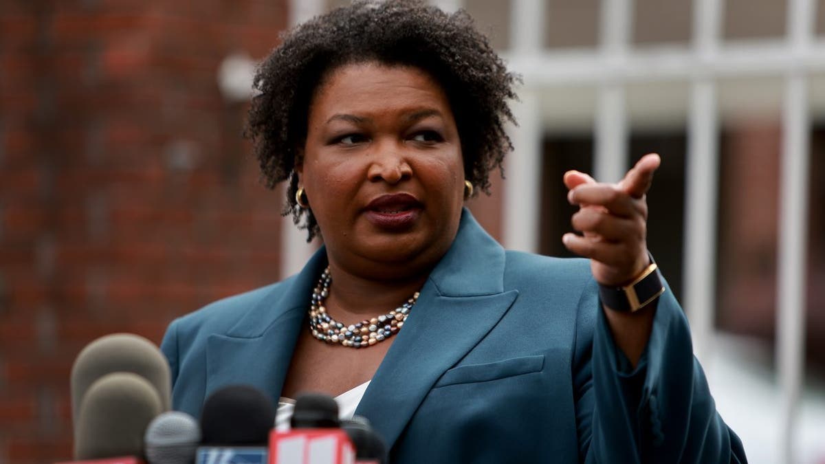 Stacey Abrams in a suit with a pearl necklace speaks in front of microphones