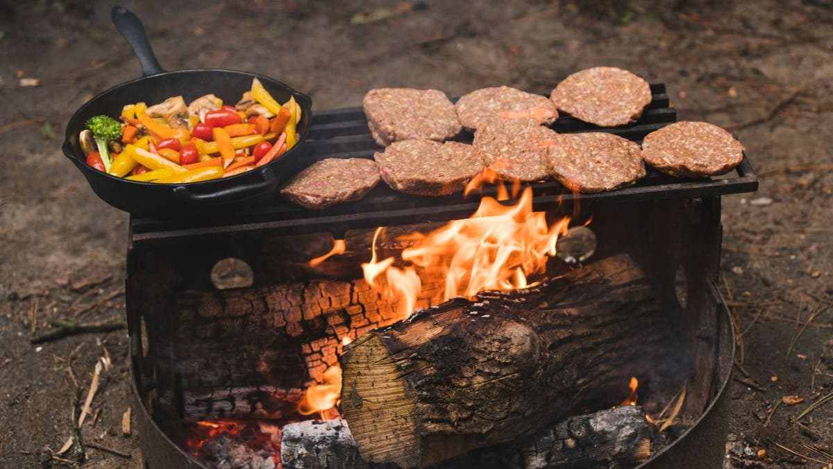 Hamburger patties rest on campfire grill next to vegetable-filled cast iron pan