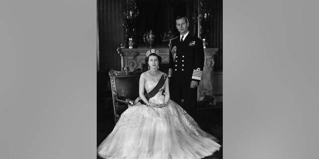 Queen Elizabeth and Prince Phillip pose for a picture once she has taken the throne. She is seated and wearing a crown, and he is standing in uniform.