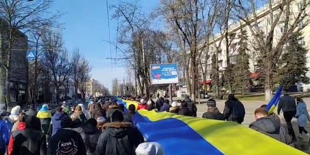 Tiešraides kadros redzams, kā cilvēki, kas 2022. gada 13. martā Hersonā, Ukrainā protestē pret Krievijas iebrukumu Ukrainā, rokās tur reklāmkarogu Ukrainas karoga krāsās, šajā nekustīgajā attēlā no sociālo mediju video, kuru ieguva aģentūra Reuters.