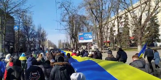 Tiešraides kadros redzams, kā cilvēki, kas 2022. gada 13. martā Hersonā, Ukrainā protestē pret Krievijas iebrukumu Ukrainā, rokās tur reklāmkarogu Ukrainas karoga krāsās, šajā nekustīgajā attēlā no sociālo mediju video, kuru ieguva aģentūra Reuters.