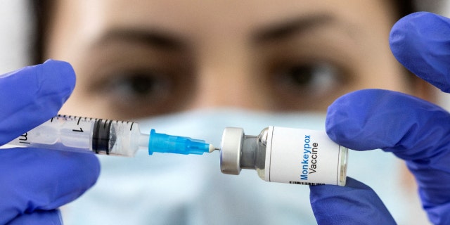 In this illustration taken on May 25, 2022, a woman holds a mock-up vial labeled ‘Monkeypox Vaccine’ and inserts a medical syringe. 