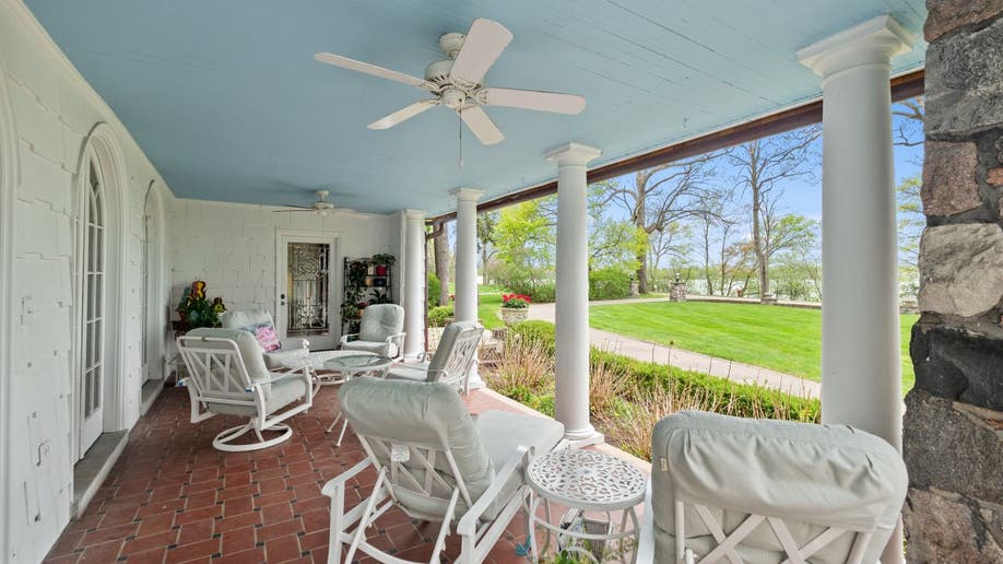 The Smurf House's front porch has three white columns and a stone wall off to the side. Furniture has been arranged for dining and lounging.