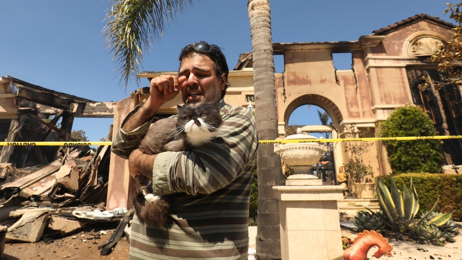A man holds his cat as he assess the damage at his home from California's Coastal Fire