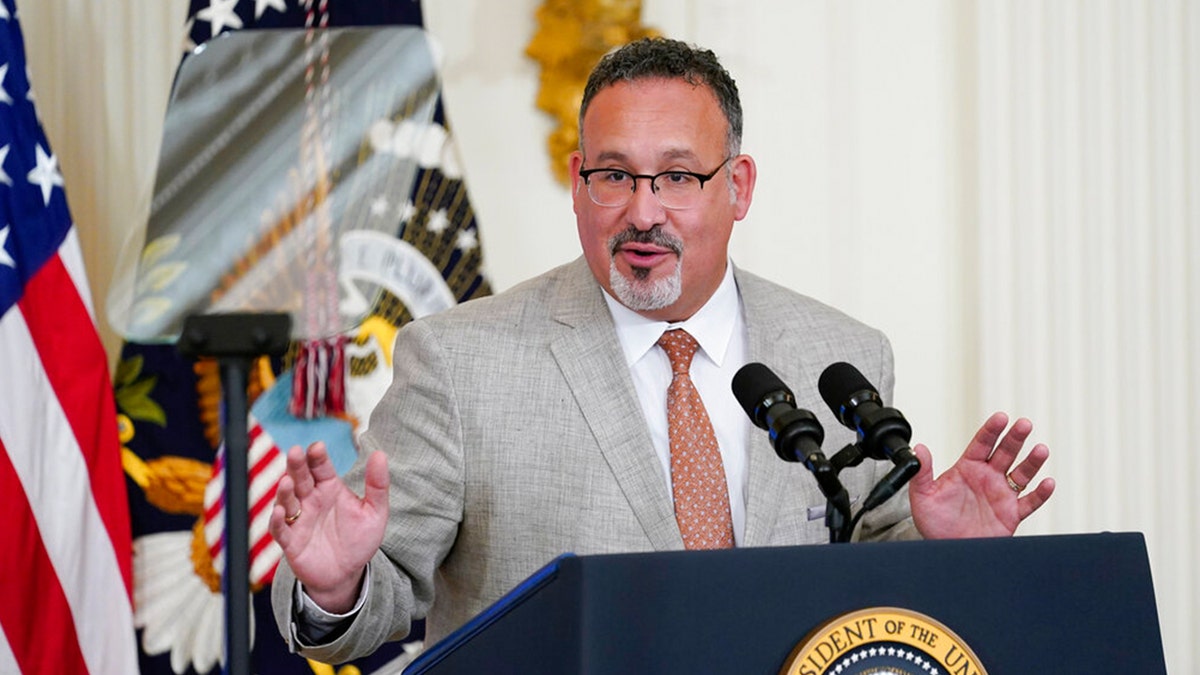 Miguel Cardona in gray suit speaks from podium with presidential seal