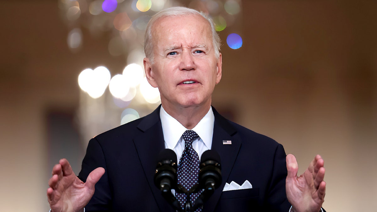 Joe Biden gestures in a black suit with blurred lights behind him