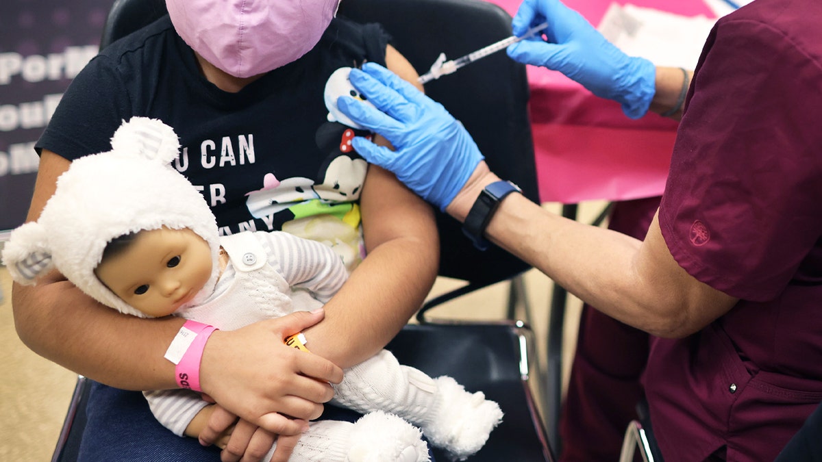 First grade student receives coronavirus vaccine while holding a doll