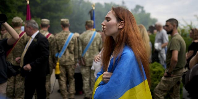 A woman wrapped in a Ukrainian flag attends the funeral of activist and soldier Roman Ratushnyi in Kyiv, Ukraine, Saturday, June 18, 2022. Ratushnyi died in a battle near Izyum, where Russian and Ukrainian troops are fighting for control of the area. 