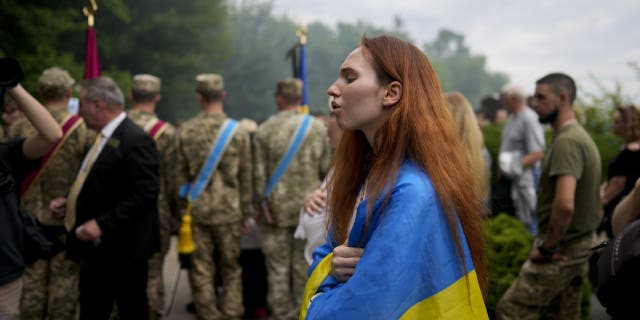 A woman wrapped in a Ukrainian flag attends the funeral of activist and soldier Roman Ratushnyi in Kyiv, Ukraine, Saturday, June 18, 2022. Ratushnyi died in a battle near Izyum, where Russian and Ukrainian troops are fighting for control of the area. 