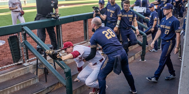 Jun 19, 2022; Omaha, NE, USA; Oklahoma Sooners first baseman Blake Robertson (26) is helped up by Notre Dame Fighting Irish pitcher John Michael Bertrand (28) after falling into the dugout during the first inning at Charles Schwab Field.