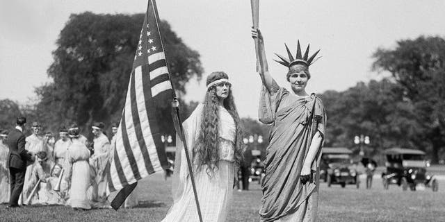 A woman holds up an American flag while wearing a white dress. Her companion wears a Statue of Liberty costume. Photo is circa 1919.