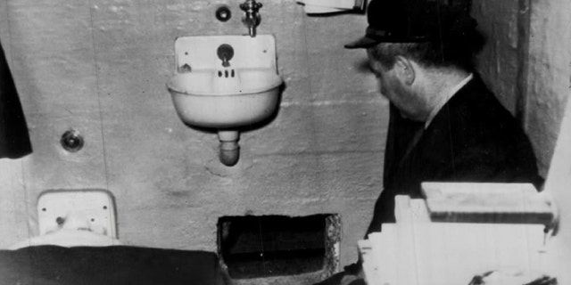 A prison guard kneels by hole in Frank Morris' cell through which he and John and Clarence Anglin escaped. Prison officials report hole was dug with broken spoons.