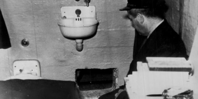 A prison guard kneels by hole in Frank Morris' cell through which he and John and Clarence Anglin escaped. Prison officials report hole was dug with broken spoons.