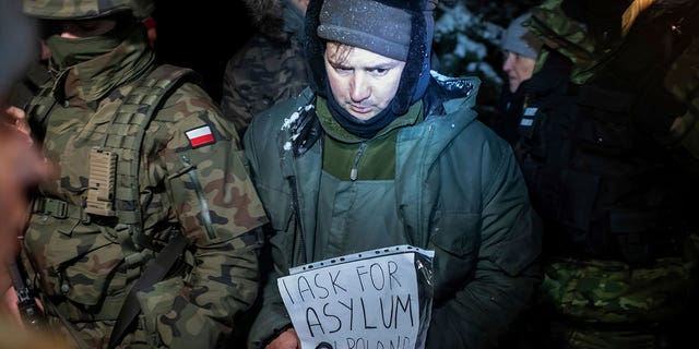 FILE - An asylum seeker from Syria who has been taken into custody by Polish border guards holds a paper saying "I ask for asylum in Poland," in Harkawicze, Poland, Dec. 1, 2021. A year after migrants started crossing into the European Union from Belarus to Poland, Polish authorities are planning to announce Thursday that a 5.5-meter-tall steel wall along its border to the north with Belarus is set to be completed. The purpose of the wall is to keep out asylum seekers fleeing conflict and poverty in the Middle East and Africa. 