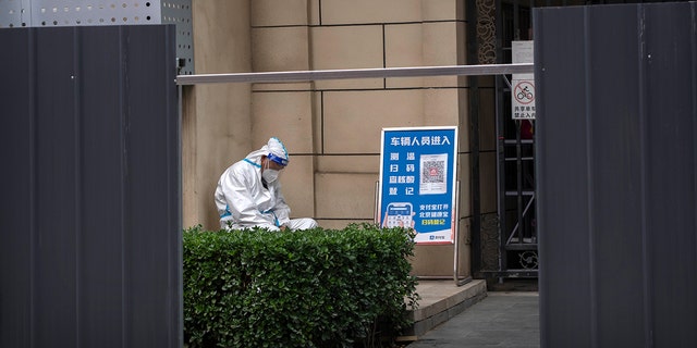 رجل يرتدي بدلة واقية يجلس عند مدخل مبنى سكني محاط بحواجز معدنية كجزء من ضوابط COVID-19 في بكين ، الثلاثاء 14 يونيو 2022.
