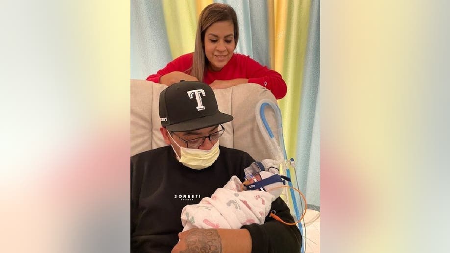 Johnny holds one of his daughters while he sits in a chair at the hospital. His girlfriend Carmen looks over the pair from behind the chair.