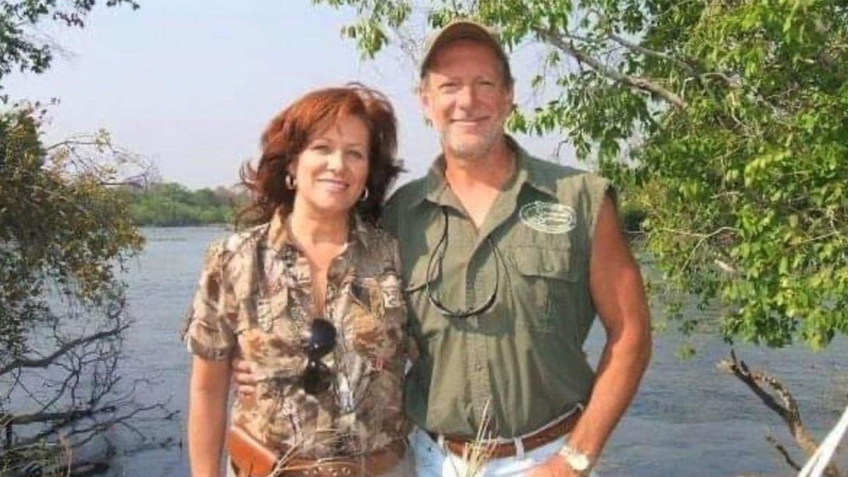 Bianca and Lawrence Rudolph pose in hunting gear in front of a lake