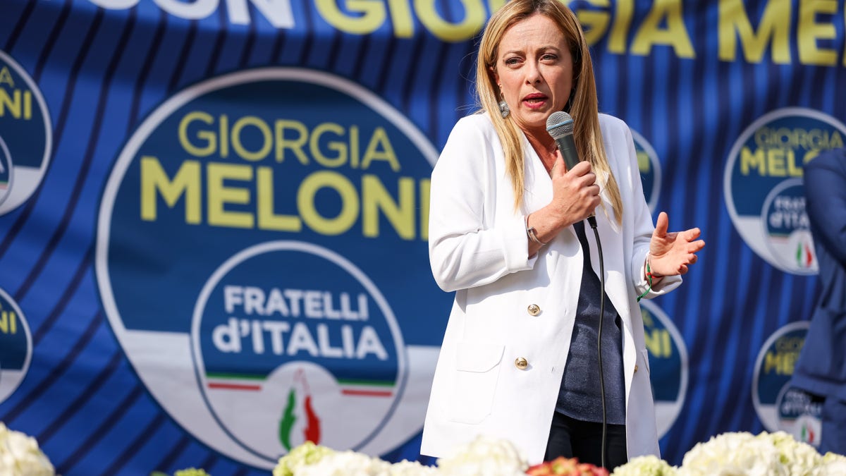 Giorgia Meloni stands in a white sport coat in front of a blue political banner