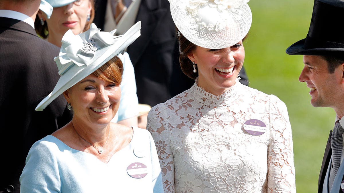 Kate Middleton smiling in a white lace floral dress and matching hat next to her mother.
