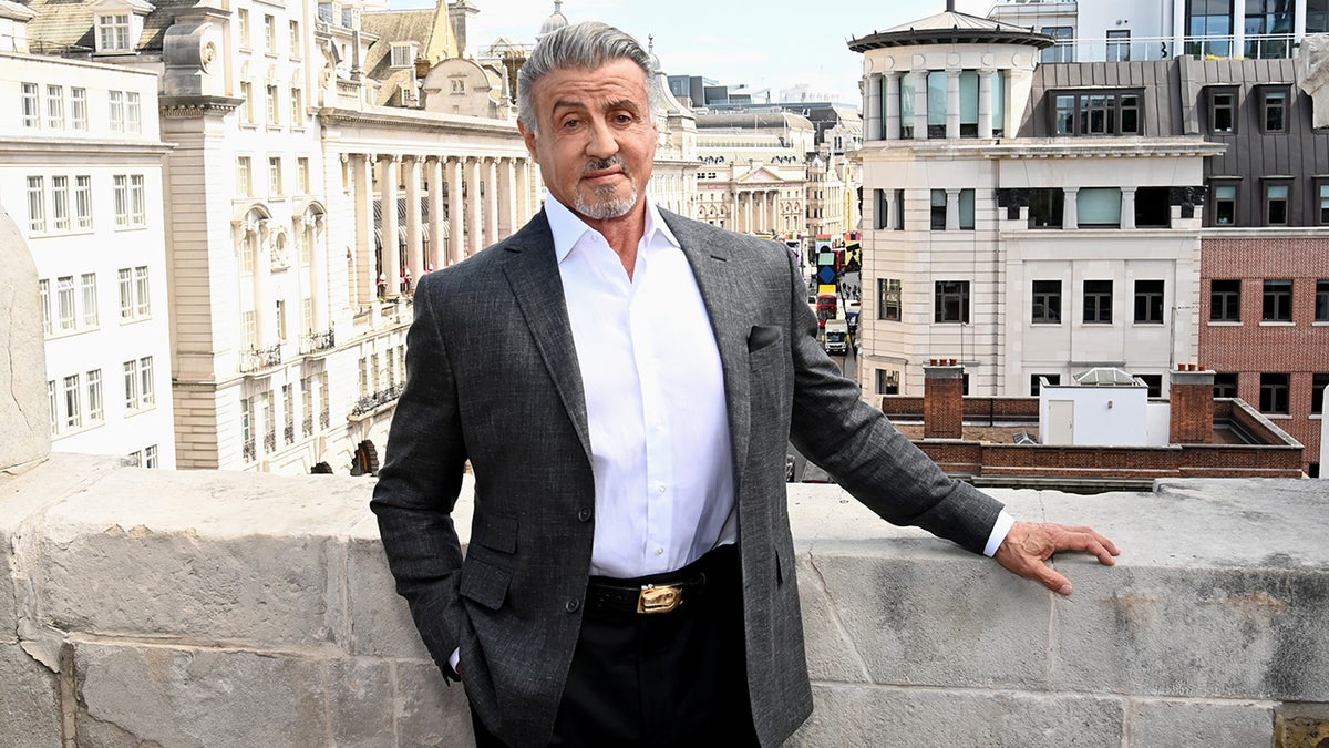 Sylvester Stallone in England poses for a photo in a white shirt and grey suit 