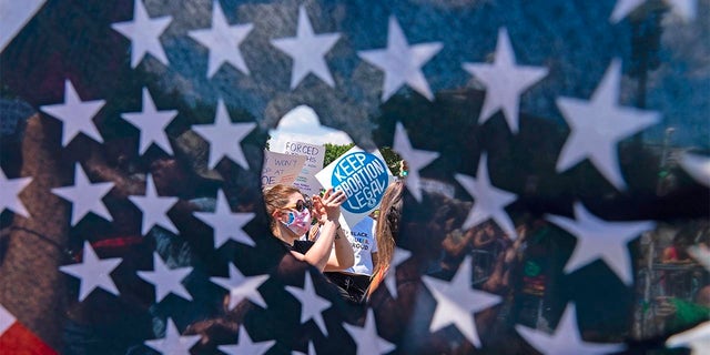 Abortion rights activists are seen through a hole in an American flag as they protest outside the Supreme Court in Washington, Saturday, June 25, 2022. The U.S. Supreme Court's decision to end constitutional protections for abortion has cleared the way for states to impose bans and restrictions on abortion — and will set off a series of legal battles.