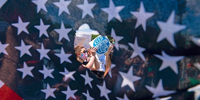 Abortion rights activists are seen through a hole in an American flag as they protest outside the Supreme Court in Washington, Saturday, June 25, 2022. The U.S. Supreme Court's decision to end constitutional protections for abortion has cleared the way for states to impose bans and restrictions on abortion — and will set off a series of legal battles.