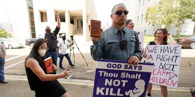 Anti-abortion activist Gabriel Olivier, center, holds his Bible and a message sign while abortion rights supporters hold counter signs and dance around him, as he and other anti-abortion supporters call out to people leaving the Hinds County Chancery Court, Tuesday, July 5, 2022, in Jackson, Miss.