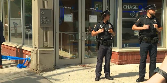 NYPD officers stand guard outside the bank's front door, with bloodstains visible on the sidewalk.