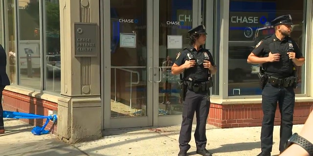 NYPD officers stand guard outside the bank's front door, with bloodstains visible on the sidewalk.